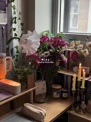 A cozy desk setup with a window view is shown. A vase of flowers is in the center of the desk, with candles and books to the right. The desk itself is made of wood, a laptop and an orange mug are to the left.