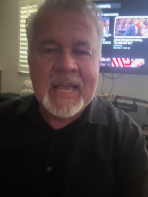 A man with gray hair and a goatee is centered, looking directly at the viewer with an open mouth. Behind him is a television with a visible screen displaying a video with text. The image has a relatively neutral indoor setting.