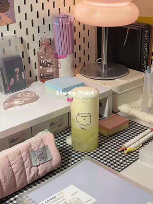 A desk setup with study supplies including sticky notes, pens and pencils, a water bottle, and a drink in a cup with a small illustration. The background has a pegboard with notes on it, and everything is set on a black and white checkered mat. The lighting is warm, coming from a lamp.