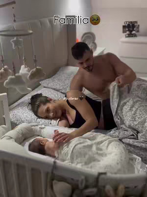 The cover of the video shows a warm and intimate family scene in a bedroom. A man, partially shirtless, leans over a bed. A woman is lying on the bed, gazing adoringly at a baby in a crib at the foot of the bed. The composition presents a moment of tenderness and care.