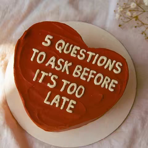 A heart-shaped cake with red frosting is centered on a white plate. White icing is used to write the text on the cake, which reads, "5 QUESTIONS TO ASK BEFORE IT'S TOO LATE."