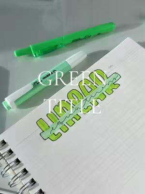 A notebook with a green spiral binding is open on a light gray surface. A green highlighter and a pen are near a handwritten title. The title is written in green marker with a quirky font. The word "GREEN" is written largely above the word "TITLE".