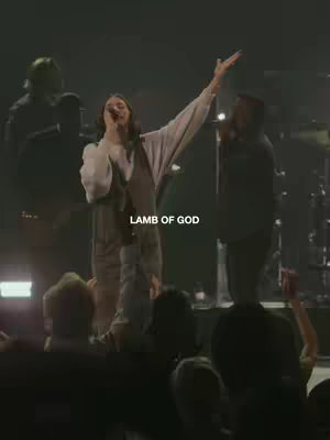 The image shows a woman on stage singing into a microphone, with her arm raised. Text overlayed says 'LAMB OF GOD'. The background is dark with stage lighting.