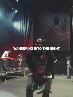 A man wearing a black t-shirt, sunglasses, and camouflage shorts stands in front of a stage with several other people performing. The image is taken from a wide angle, showing the stage and audience.