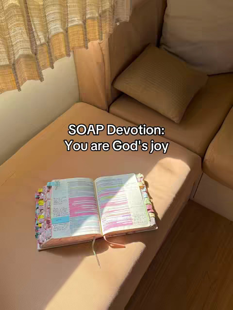 An open Bible lies on a light brown couch, with colorful tabs and notes, a window with beige curtains is on the left and a pillow is to the right