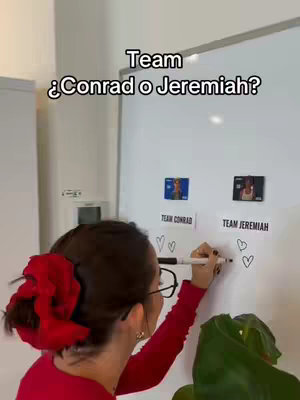 A person is standing in front of a whiteboard where two small images are placed. The whiteboard is labeled with "Team ¿Conrad o Jeremiah?" The person is holding a black marker and drawing hearts on the board. The scene is well-lit, and the background includes a neutral-colored wall and a green plant.