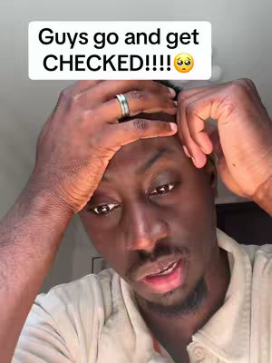 A selfie-style shot featuring a Black man. He is holding his hands to his head, looking downward with a concerned expression. A text overlay at the top reads, "Guys go and get CHECKED!!!!" with a worried face emoji.