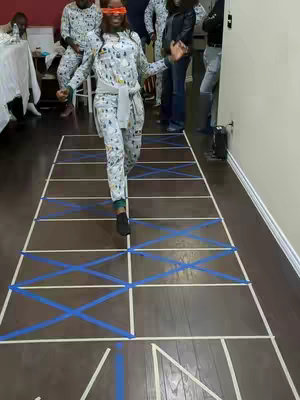 The image shows a woman wearing a jumpsuit and an orange blindfold, attempting to walk on a path of taped squares on the wooden floor. Other people in matching pajamas are in the background.