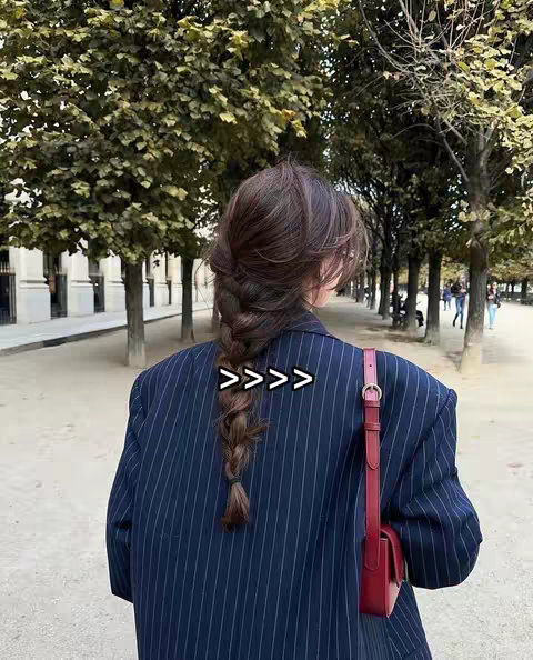 A woman with long brown hair styled in a braid is seen from the back. She is wearing a navy blue pinstripe blazer and carrying a red purse over her shoulder. The background features a blurred street scene.