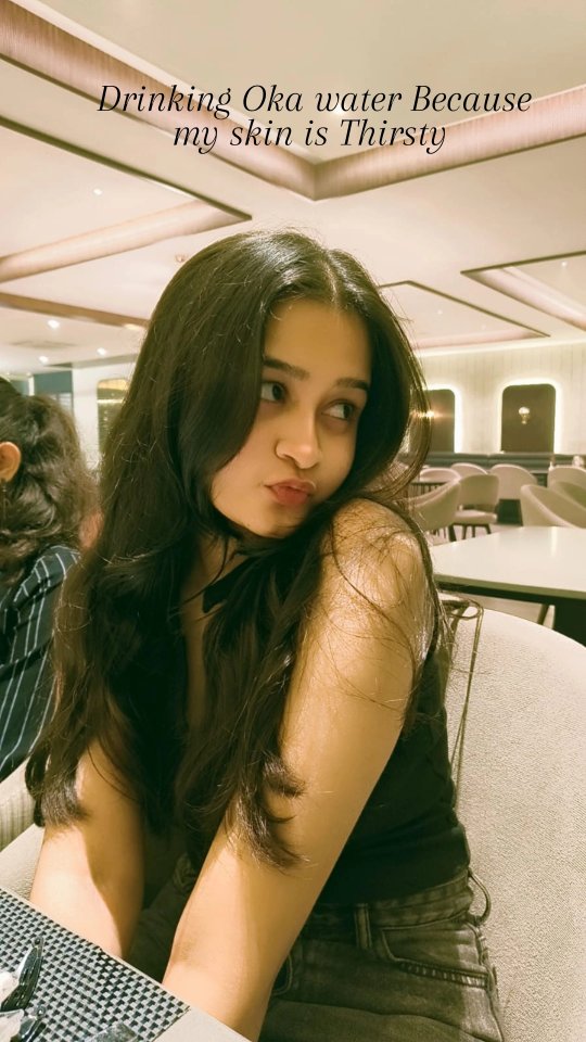 A young woman is looking toward the right, with slightly pursed lips as she makes a kissing face. She has long, dark, wavy hair. She is seated in what appears to be a restaurant, with a blurred background of tables and lighting.