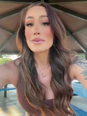 A selfie-style shot of a woman from the chest up, posing under a gazebo-like structure. She has long, wavy brown hair and is wearing a brown top and a necklace with a pendant. The background shows a park setting with blue benches; the lighting seems natural, with the underside of the gazebo casting slight shadows.