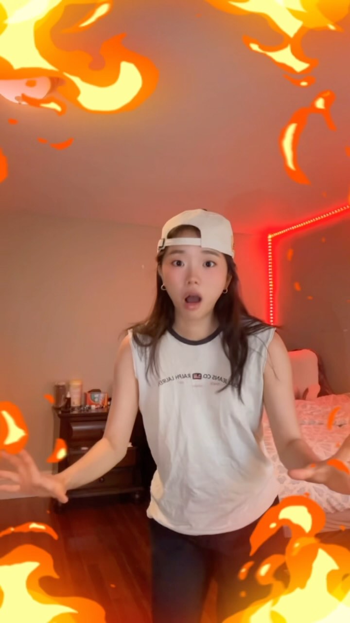 A young woman with wide eyes and an open mouth is framed by cartoon flames, creating an excited or surprised expression. She is wearing a white baseball cap and a sleeveless white t-shirt. The background is her bedroom with a red light from a light strip.