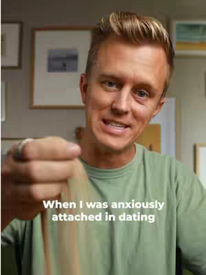 A man with light skin and blonde hair is looking directly at the camera in a well-lit interior setting. The man is wearing a light green t-shirt and has a light brown substance in his hand, with his left hand holding the material. He's smiling slightly. Behind him, the background wall displays framed artwork and various other pictures. Overlaying the image is white text at the bottom stating, 'When I was anxiously attached in dating'.