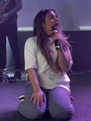 A woman is kneeling on a dark purple stage, holding a microphone up to her mouth as if she is singing or speaking with great emotion. She is wearing a white t-shirt and light-wash jeans. The stage presence suggests a performance or a public speaking event.
