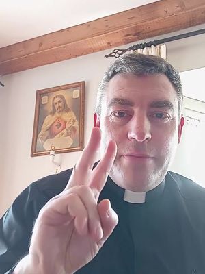 A man in a priest's collar is taking a selfie. He is holding up a peace sign with two fingers. Behind him is a painting of Jesus and a wood-paneled ceiling.