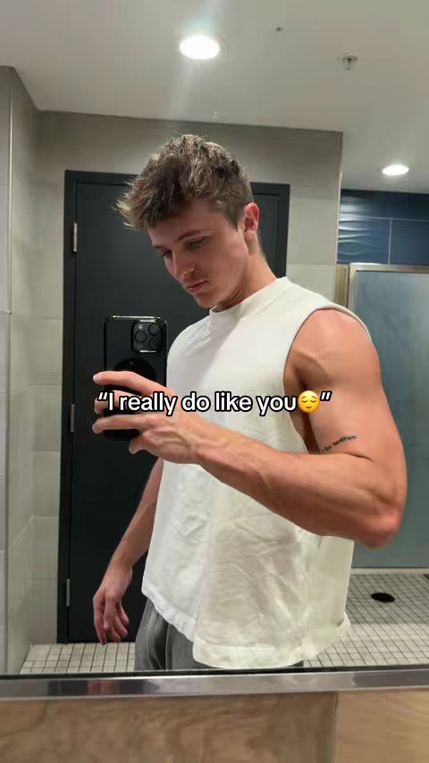 A man in a white tank top is taking a mirror selfie in what appears to be a bathroom. The background includes a closed black door and a tiled wall. Text overlayed on the image reads: 'I really do like you' with a smiling face emoji.