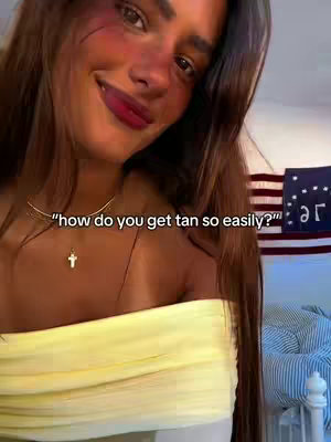 A smiling young woman with long brown hair poses for a selfie. She is wearing a yellow tube top and a necklace with a small pendant. The background includes a striped flag and a bed.