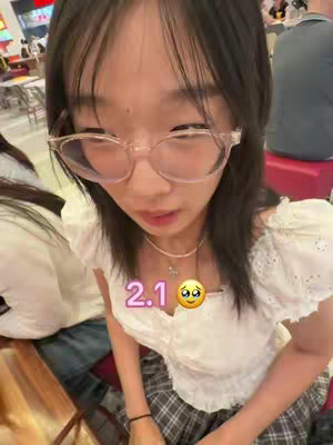 A young woman with glasses looks down with a slightly concerned expression. The image has a close-up perspective focusing from her chest up. She is wearing a white top and is seated in a public place. The lighting is natural and bright.