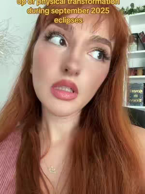 A close-up shot of a woman with long red hair. The woman has fair skin, large eyes with visible eyelashes, and full lips. Her expression is one of surprise or slight concern, with her mouth slightly open. She wears a rose-colored top and a gold necklace. The background is slightly blurred, but appears to be a light-colored wall with some shelves and decor.
