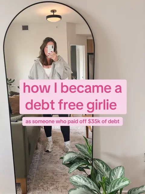 A woman is reflected in a large, arched mirror, holding up her phone to take a selfie. She is wearing a gray hoodie and leggings, standing in front of a green couch and a houseplant. Pink text boxes overlay the image, stating "how I became a debt free girlie" and "as someone who paid off $35k of debt."