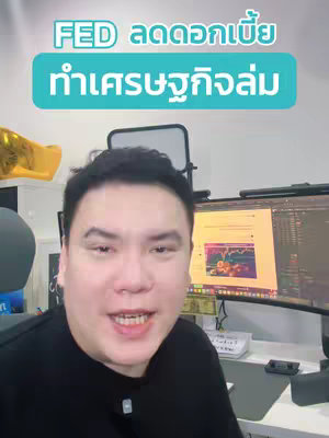 A man with dark hair and a black shirt is the focus. Overlayed on the image is a turquoise rectangle with white text, 'FED ลดดอกเบี้ย ทำเศรษฐกิจล่ม'. He is framed from the chest up, in front of a screen showing charts and graphs. An inflatable gold figure is in the background.