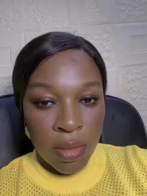 A close-up shot of a person with dark skin, staring at the camera. The person is wearing a yellow top with a mesh design. The background is a patterned wall. The lighting illuminates the face.