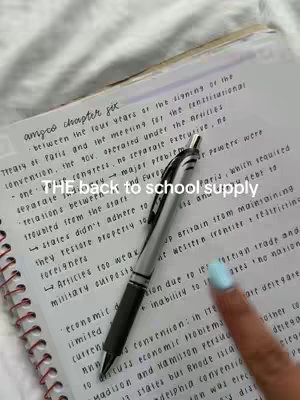 An overhead shot featuring a notebook open to a lined page filled with handwritten notes. A black and silver pen is placed diagonally across the open notebook pages. A finger with a light blue painted nail is visible in the bottom right corner.