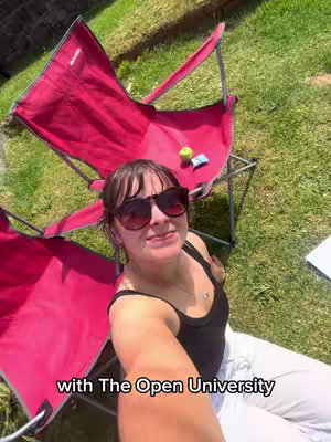 A woman wearing sunglasses is smiling, sitting in a red folding chair in a grassy yard. Another red chair is behind her, and a green apple and a small packet are on the chair behind. Text at the bottom says 'with The Open University'.