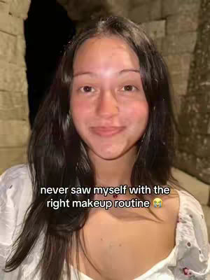 A selfie-style shot of a young woman with dark hair, looking at the camera with a slight smile. She is in an outdoor setting, with a stone wall as the background. The text overlay reads, 'never saw myself with the right makeup routine.'