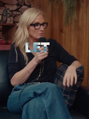A woman with blonde hair and glasses speaks into a microphone while seated. She is wearing a black long-sleeved shirt and jeans. The background is a mix of wooden paneling and what appears to be hanging plants.