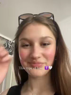 A young woman with fair skin, is smiling and looking towards the camera. She has sunglasses perched on her head. On her cheeks, she has a rosy blush. The video overlay has the text 'This type of love' with a religious symbol and a heart emoji.