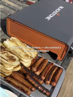The image shows a Blackstone griddle with cooked sausages and sliced grilled bread. The griddle is on an outdoor surface, suggesting a cookout setting. Text overlay indicates the item may be a gift.