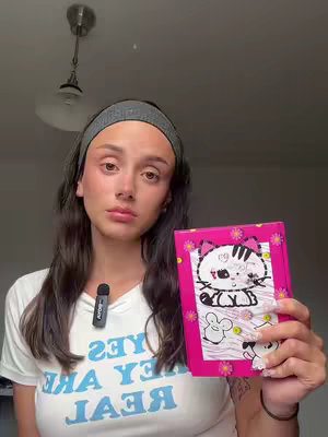 A young woman with long dark hair, a gray headband, and light makeup is holding a pink journal featuring a cartoon cat, in front of a simple light-colored wall and a dark, low-hanging light fixture, with an attached, small microphone.