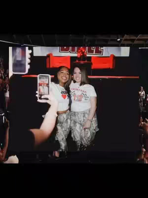 Two women take a selfie on a stage or event, with a banner in the background that says 'BIBLE'. The woman on the left is holding the phone, partially obscuring her hand. The woman on the right has her arm around the woman on the left. They appear to be smiling, with the background out of focus with a large crowd.