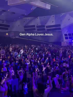 A large indoor space is shown, likely a large hall or auditorium, filled with a crowd of people. The ceiling is high, and the lighting is a mix of cool-toned blues and purples, creating a dynamic atmosphere. The image is filled with the silhouettes of many people, with some raising arms, suggesting music or an event is taking place. The composition is crowded, reflecting the energy of a live gathering.