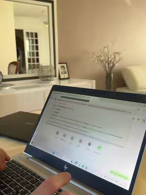 A shot is taken from the point of view of someone working on a laptop. The screen is open displaying a browser-like interface. Foreground consists of the laptop, user’s hand and fingers, and a secondary laptop. Behind the laptops are a white surface with decorations and a mirror. The lighting is natural.