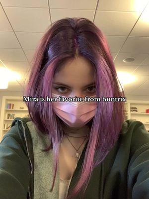 A selfie-style shot featuring a young woman with pink and purple hair, wearing a pink face mask and a green hoodie. Text overlaid reads "Mira is her favorite from huntrix". The background shows bookshelves.
