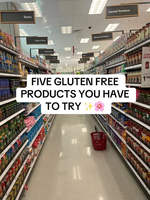 A wide-angle shot inside a grocery store aisle. The aisle leads toward the back of the store, with shelves on both sides stocked with various food products. Above, signs identify the sections: "Beans," "Sugar & Flour," "Macaroni & Cheese," and "Canned Tomatoes." In the foreground, a shopping basket sits on the floor. Text overlays in the center read: "FIVE GLUTEN FREE PRODUCTS YOU HAVE TO TRY" with a sparkle and flower emoji.