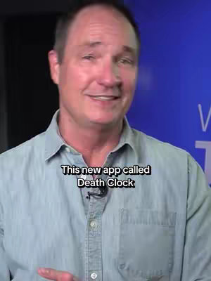 A middle-aged man in a light blue button-down shirt stands in front of a blue background. A text overlay in a black box with white letters reads, 'This new app called Death Clock'. The man is smiling and looking towards the camera, with a microphone visible on his shirt.