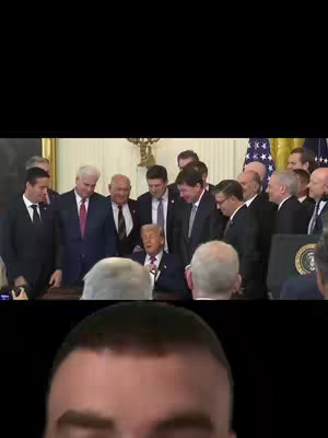 The cover image is split into two main sections: the top portion features a video screen displaying a group of men in suits surrounding a seated man signing documents, likely in an official setting. The lower section shows a close-up of a man's face. Both sections are framed by a black border.