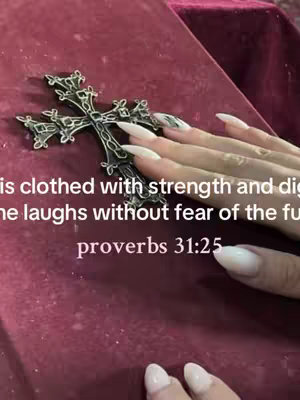 The image displays a hand with long, manicured nails holding a decorative silver cross. The cross is positioned over a textured, burgundy surface that appears to be fabric. Text is overlaid in white font, with the verse from Proverbs 31:25