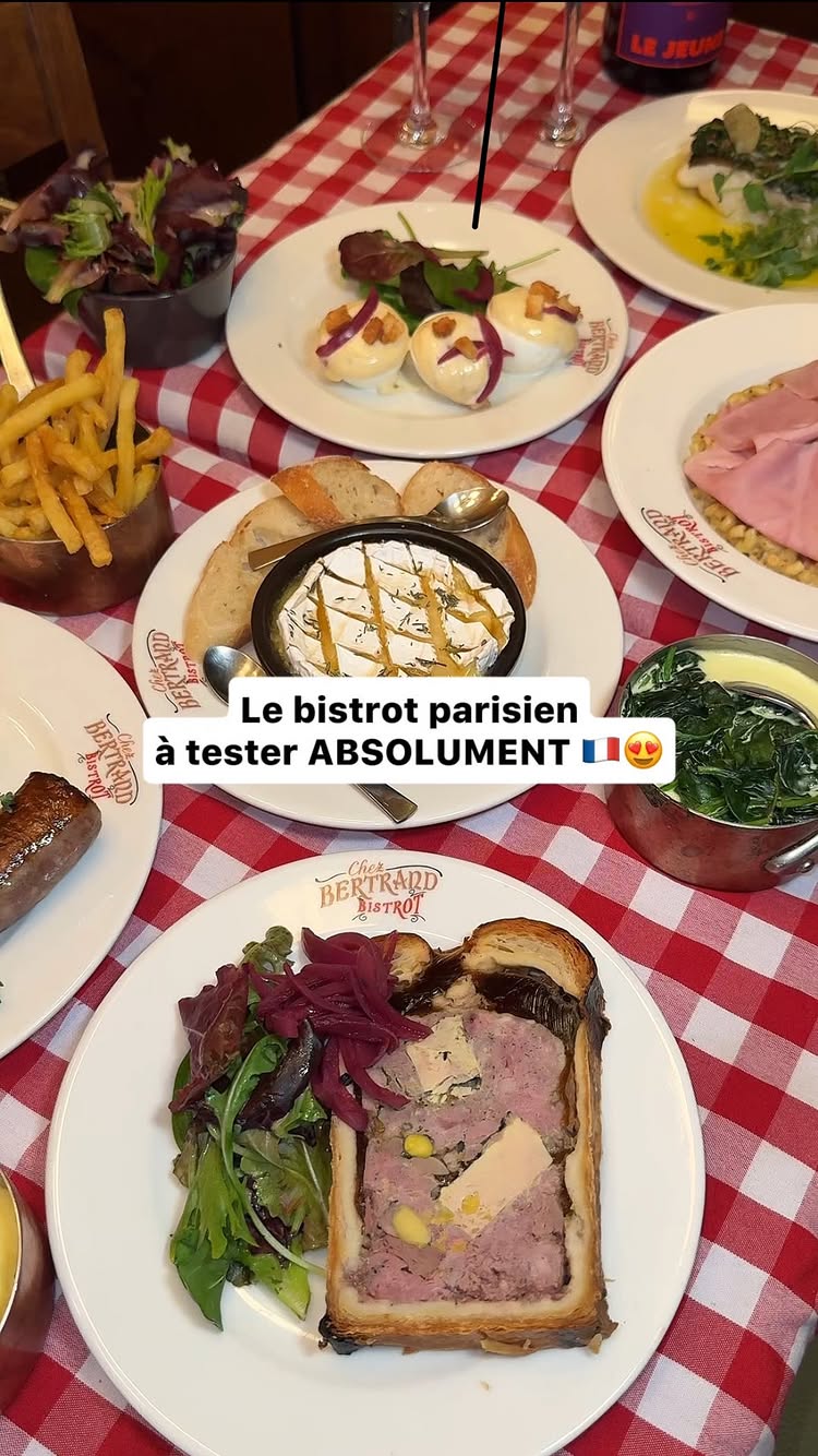 The image showcases a table laden with various dishes, presented in a slightly over-the-shoulder angle. The dominant color is a bright, checkered red and white, forming the tablecloth’s pattern. Plates of food occupy most of the frame, including what appears to be a starter of fries, a pot of spinach, a sausage, and a pate; the centerpiece is a bowl with bread. The text overlay in the image suggests a bistro.