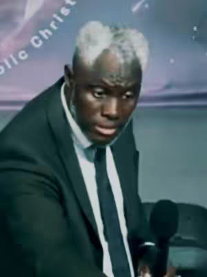A man dressed in a suit with a black tie and white collar is seen in a medium shot. He is holding a microphone and looking down with a slight pensive expression. The background has a blurred, light purple gradient with white text.