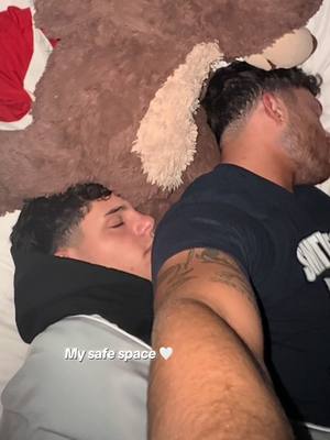 The image features two young men asleep in bed, embracing. One is partially visible with his head nestled against the other figure's chest. The background includes a portion of a large brown teddy bear. Text overlay reads "My safe space" with a white heart symbol.