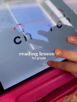 The image shows a hand touching a tablet that is displaying a reading lesson. White text overlays the image reading "reading lesson" and "1st grade". There is a pink surface visible below the tablet.