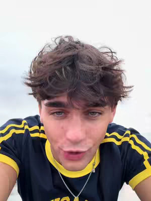 A selfie-style shot featuring a young man, framed from the chest up, taken with a slight upward angle. The background is a washed-out, light-colored surface, possibly the sky or a bright wall, giving a sense of soft lighting. The man has tousled dark hair and is wearing a dark blue t-shirt with yellow accents along the neck and shoulder seams. He's looking directly at the camera with a slight smile, and a silver necklace hangs around his neck.