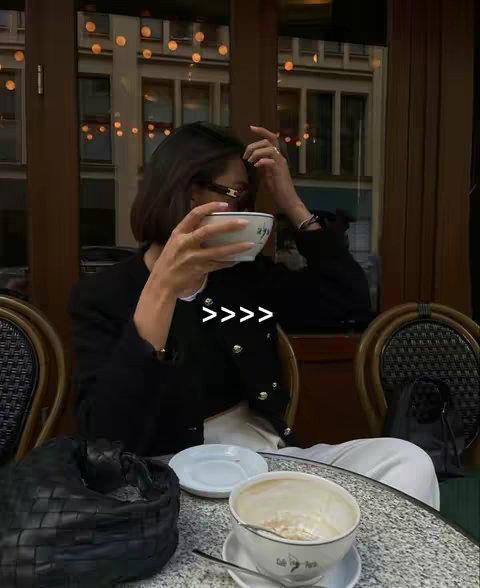 A person is sitting at a cafe table, holding a white cup with two words on it. The person is seen from the shoulders upwards. In the background, there are blurred buildings and a window. The person is wearing a dark-colored coat or jacket. The text overlay reads: >>>>