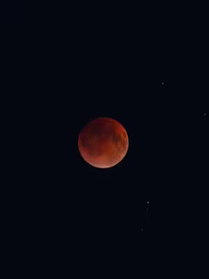 A photograph of the night sky dominated by a large, round, reddish-orange moon, with a deep black background. A few small, faint stars are scattered around the moon.