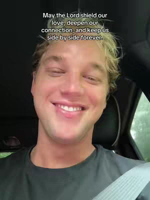 A close-up shot of a smiling man inside a vehicle, likely a car. He's wearing a dark shirt and a seatbelt is visible. Above him, overlaid text in white font. The background is blurry, showing a mix of interior and exterior details.