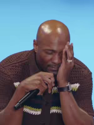 A man with a shaved head rests his head on his left hand in a moment of introspection. He wears a brown, textured knit shirt with a white and green trim, and a watch with a black strap. His right hand is held in a prayer-like position, showing a ring on his finger. The background is a solid, light blue.
